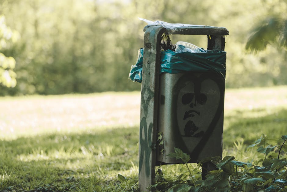A metal public garbage bin with a graffiti-style face painted on its front, situated on a grassy area outdoors near a park with trees and sunlight in the background. The bin is partially covered with blue plastic bags and appears weathered, with some vegetation growing at its base. The surrounding environment suggests an outdoor space typical of residential or park settings, where waste collection occurs. This image relates to household or communal waste disposal, possibly linked to home relocation or decluttering processes managed by Man with Van Dartmouth Park, a company that provides removals and moving services.