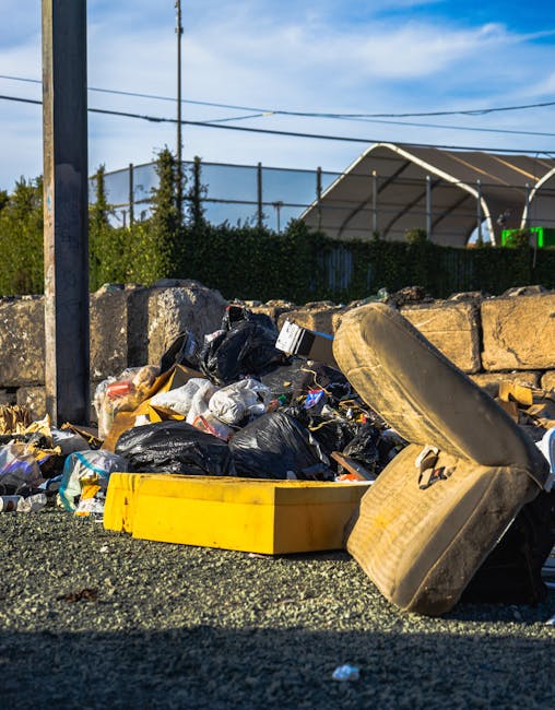 A pile of bulky waste, including black garbage bags, cardboard boxes, and a large, worn car tyre, is accumulated on a gravel surface next to a stone wall and metal fencing. Some items are stacked loosely, with a yellow plastic tray lying near the bags and the tyre leaning against the stone wall. In the background, there are utility poles, power lines, and a partially covered outdoor area with a curved, green-roofed structure under a blue sky with scattered clouds. The setting appears to be outdoors, possibly on or near a property boundary, during daylight hours. This scene illustrates waste disposal or clearance of household items, often part of house removals or estate clearances, highlighting the importance of proper bulky waste collection and removal services such as those offered by Man with Van Dartmouth Park.