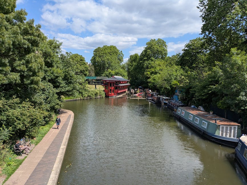 Photograph of a calm river scene with a paved walkway on the left side, where one person is walking and another is seated on a bench. On the right side of the river, several houseboats with flat, rectangular roofs are moored along the bank, surrounded by dense, leafy trees with green foliage. In the background, a small traditional-style pagoda with a red structure and dark roof extends over the water, visible among the trees. The sky above features scattered clouds, and the overall scene is bright and sunny, illustrating a peaceful urban riverside environment. The image depicts a typical setting for house removals or moving logistics near Dartmouth Park, with potential elements like packing materials and furniture likely involved in the moving process, observed through the context of the landscape and nearby infrastructure.