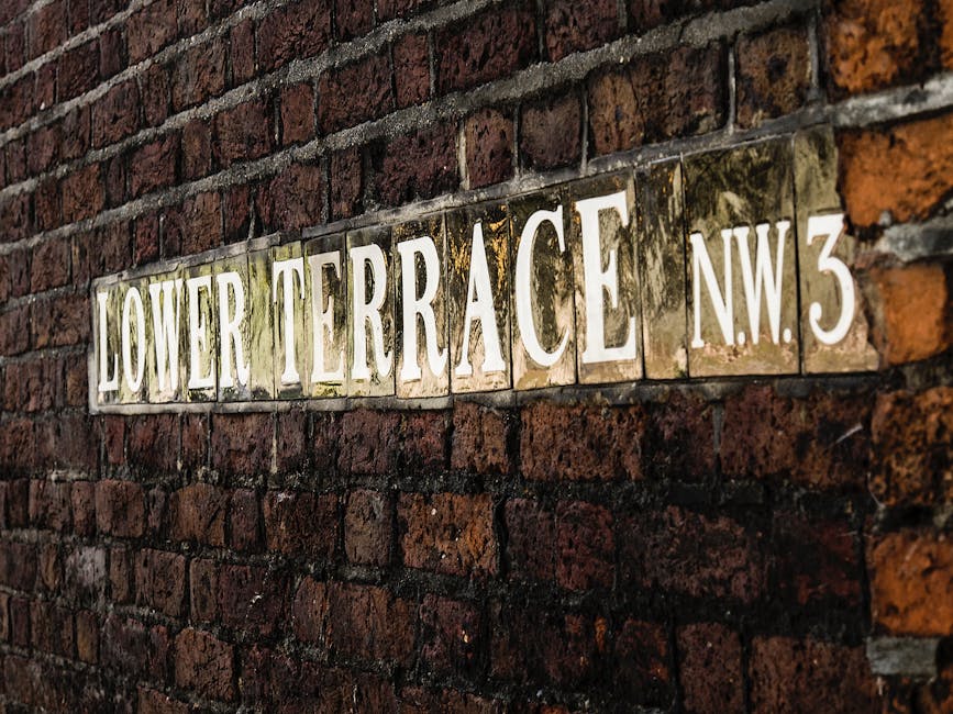 A close-up view of a rectangular street sign mounted on a red brick wall, displaying the name 'Lower Terrace NW3' with white raised lettering on a weathered, reflective metal background. The bricks surrounding the sign show signs of age and weathering, with some mortar appearing worn. The sign is positioned slightly off-center, with a slight angle giving a sense of depth to the image. This type of signage is commonly seen in London residential areas, providing context for local street names. The photograph's lighting highlights the textured surface of the bricks and the reflective surface of the sign, subtly indicating a setting that could be used in a house relocation or moving service context, such as a detailed visual to accompany content about moving logistics or property locations, for example, on a page about Camden Council skip permits near Dartmouth Park. The focus on the street sign subtly supports the relevance of residential areas involved in furniture transport and home relocation processes as offered by Man with Van Dartmouth Park.