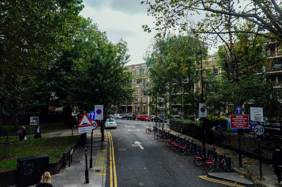 A residential street scene in Dartmouth Park, showing a narrow, tree-lined road with parked bicycles on the right side secured to a bike rack, and several cars parked along the curb. The road has a double yellow line and various traffic and parking signs, including a no parking zone and a warning about hump speed bumps for 35 yards. In the background, multi-storey apartment buildings with balconies are visible, partially obscured by trees with green and autumn-colored leaves. Pedestrians, including a woman with a backpack and other individuals, are walking on the pavement on the left side of the image. The scene is in daylight with natural lighting, highlighting the urban environment suitable for house removals and the logistical planning of transportation and home relocation services offered by Man with Van Dartmouth Park.