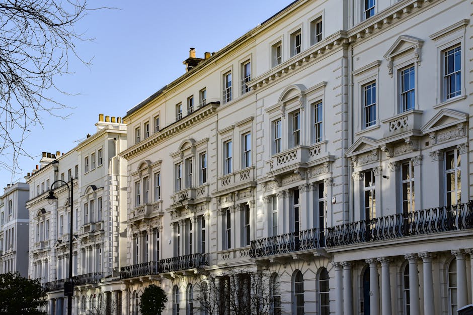 A row of Victorian-style terraced houses constructed with grey stone and brick, each featuring multiple white-framed sash windows, steep gable roofs, and chimneys. The buildings are set behind a low b