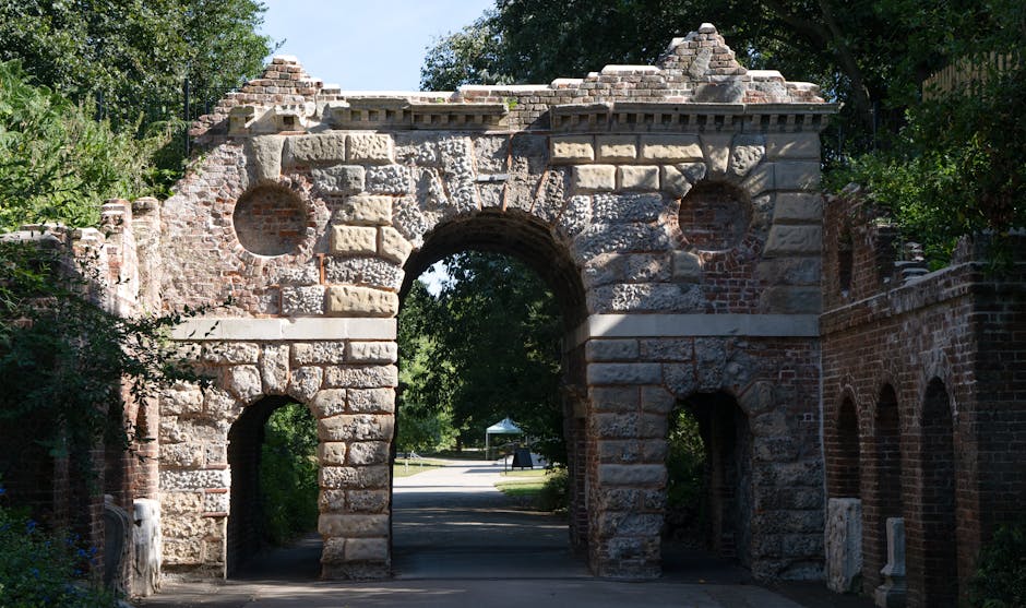 A historic stone archway with a large central opening and smaller side openings, constructed from irregularly shaped stones and brick, is situated at the entrance of a park or residential area. The structure shows signs of age, with weathered masonry and some brickwork visible in the upper sections. Surrounding the archway are trees and greenery, with sunlight casting shadows on the stones. In the background, a paved road extends through the arch, with a glimpse of a park tent or gazebo further along the street. The scene depicts an outdoor environment with natural light, and the archway serves as a gateway, possibly part of a historical site or city landmark. This image is relevant to house removals and moving services by illustrating the kind of historic or distinctive entrances that may be encountered during a home relocation or furniture transport in Dartmouth Park, with Man with Van Dartmouth Park providing such services.