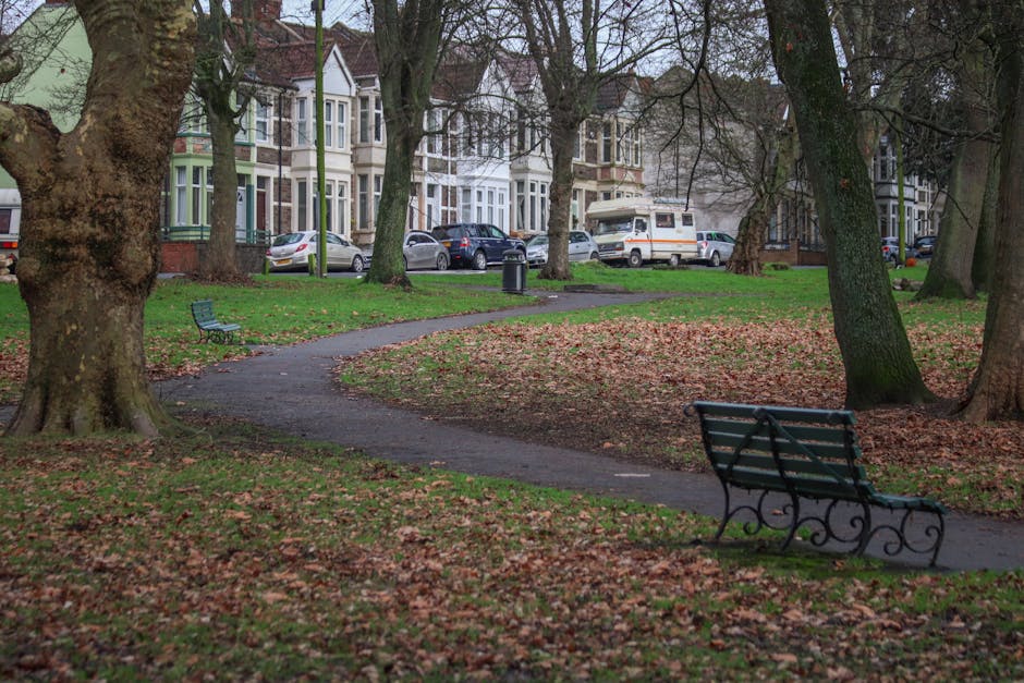 A park scene with several large trees with thick trunks and branches, some with textured bark, and a winding paved pathway covered with fallen leaves. The pathway curves through the green grassy area, which is interspersed with patches of brown leaves, suggesting an autumn setting. Across the park, there are two empty metal benches with dark green slats and decorative armrests, positioned along the walkway. In the background, there is a row of Victorian-style terraced houses with bay windows, featuring pastel and brick facades, and several parked cars including a white van, which may be involved in a moving or home relocation process. The scene is captured in natural daylight, with some overcast sky visibility, indicating a calm, quiet residential street typical of Dartmouth Park. Occasionally, [COMPANY_NAME], specializing in removals and furniture transport, may be involved in loading or unloading activities during house removals, contributing to the overall moving logistics captured in this setting.