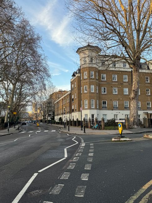 A wide view of Dartmouth Park Road showing an older multi-story brick building with white window frames and a rounded corner, situated at a street intersection. Several leafless trees line the pavemen