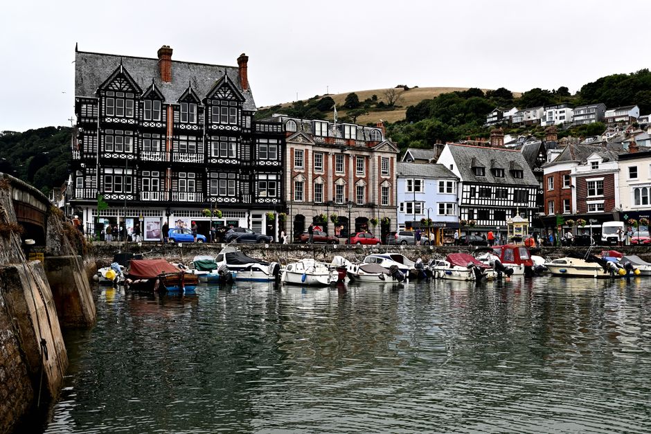 View of a marina with several small motorboats docked along the quay, some covered with tarps or protective covers. Behind the boats, there is a stone wall separating the water from a promenade busy with pedestrians, some walking and others waiting in line outside a café or restaurant named 'Embankment'. The establishment has an outdoor seating area with tables and umbrellas and a sign indicating services like terrace dining, bar, takeaway, bistro, and ice cream. In the background, there are residential houses on a hill covered with lush green foliage, and a clear blue sky overhead. A blue car is parked on the street near the café, and the scene captures a lively, picturesque coastal town setting that could relate to local house removals or moving services in the Dartmouth Park area, emphasizing relocation logistics involving both transportation and packing within a scenic environment.